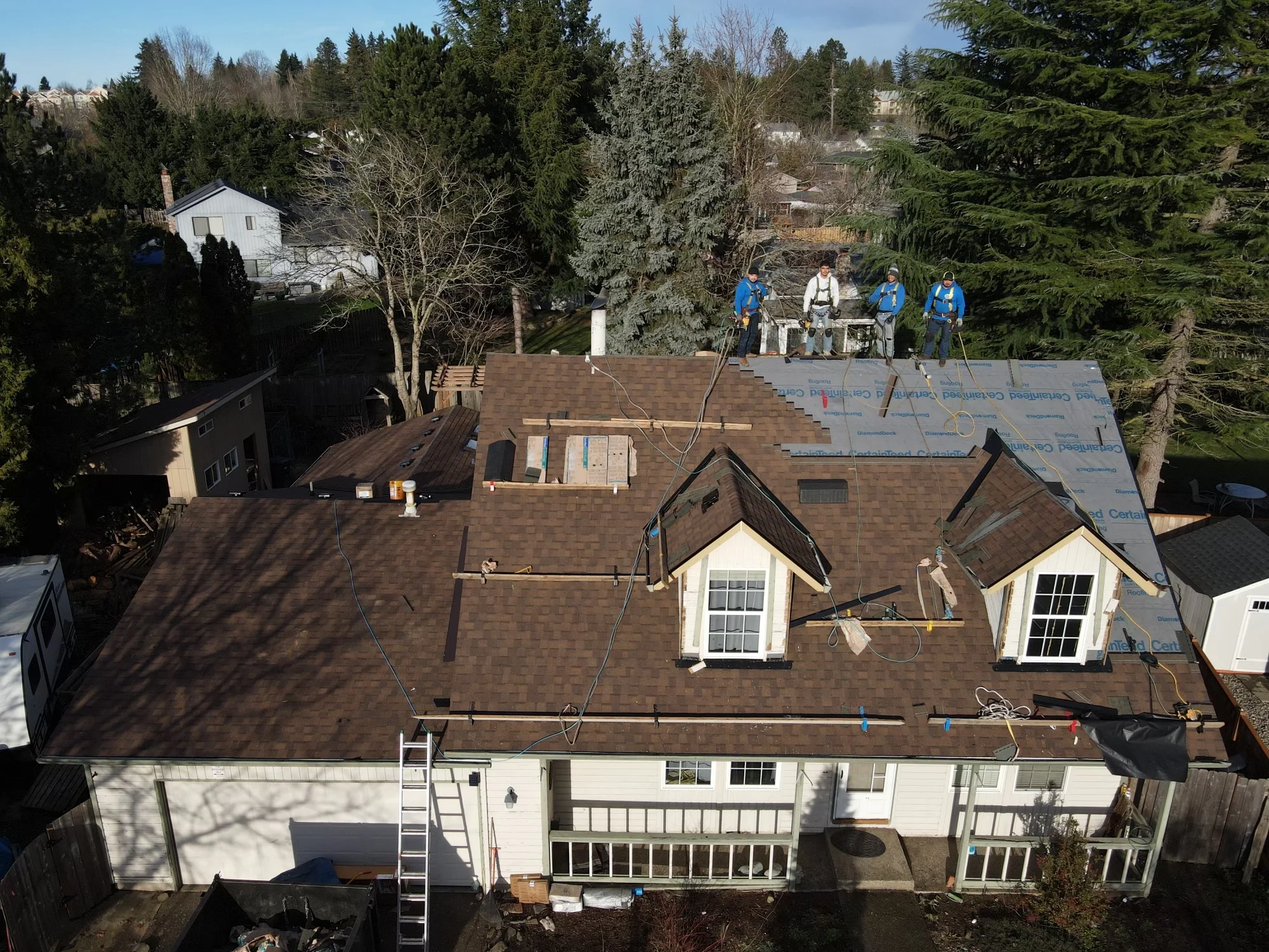 Roofers installing brown architectural shingles on residential home with dormers and white trim
